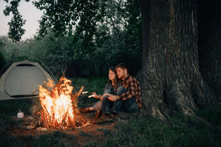 lovers man and woman sit by the fire and fry marshmallows. Camping in the forest. Enamored guy and girl travelers are preparing food. Outdoor tourism and recreation concept.の写真素材