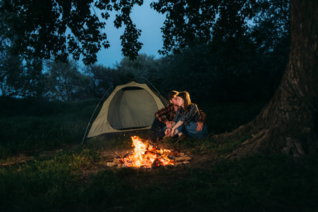 traveler couple in the forest resting by the fire after a working day. Man and woman students are sitting near the tent. Trekking and tourism concept. Autumn and evening.の写真素材