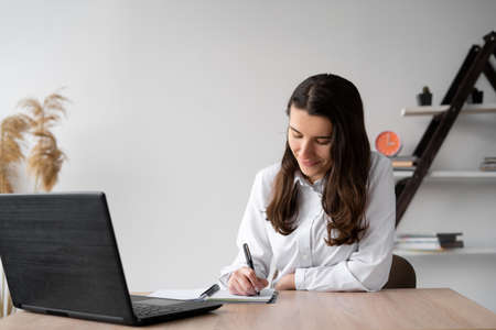 Young girl student studies at home online with a laptop while writing homework in a notebook while sitting at a desk. Remote work or education. lesson or conference via a laptop web.の写真素材