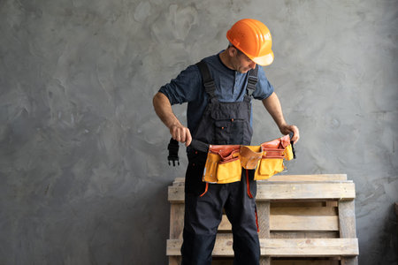 A male builder or repairman puts on a tool belt. The constructor stands against a gray wall wearing...