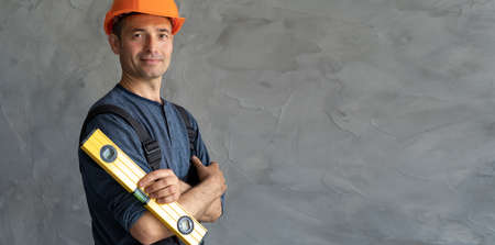 smiling construction foreman standing against gray background holding level near concrete wall. Repairman full length safety helmet wearing blue coveralls.の写真素材