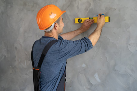 Rear view of a foreman in a helmet measuring the wall with a level. A construction worker checks the...