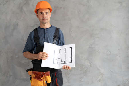 an engineer in a helmet at a construction site near a concrete wall stands with blueprints. Builder with a plan and drawings inside the house. Renovation and architecture concept.の写真素材