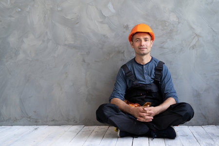 a builder sitting on the floor in a lotus position is resting from work at a construction site. A repairman or foreman sits near a concrete wall wearing a hard hat near a space for text.の写真素材