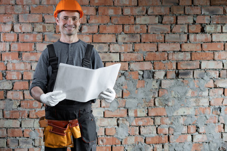 smiling builder with a plan or drawing in his hands standing near a brick wall in the house. An...