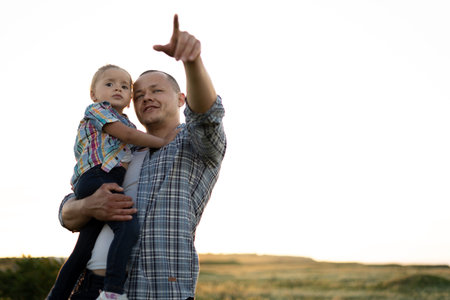 a young father holds a little daughter in his arms and points a finger to the sky in summer in...