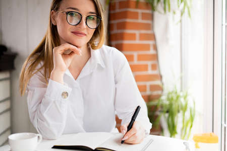 portrait of a smiling girl working in the morning in a cafe making notes in a notebook or planning the day. Close-up of a blond woman wearing glasses. Positive employee take a break.の写真素材