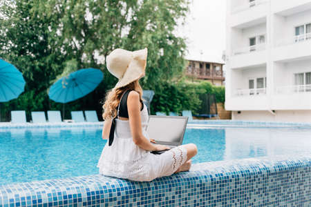 At a resort hotel near the pool sits a young happy woman in a straw hat with a laptop. technology concept and remote work.の写真素材