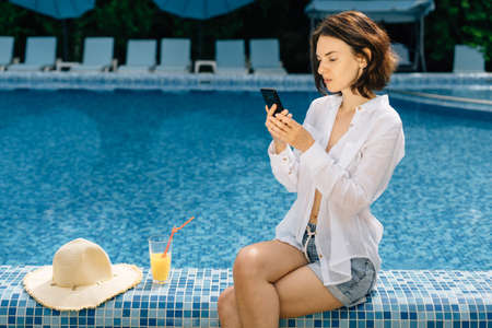 girl with a tropical cocktail or orange juice sitting on the edge of the hotels swimming pool chatting online or using social networks on her mobile phone. Recreation concept.の写真素材