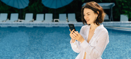 cheerful woman having a rest near swimming pool with a mobile phone leads a live broadcast or has a...