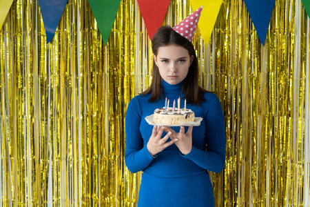 A sad woman on a yellow background in a party hat stands and holds a cake in her hands. Birthday...