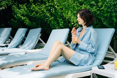 a lonely serious girl is resting in a hotel sitting on a chaise-lounge drinking juice from a glass with bare legs thinking about life. vacation concept. Lifestyle and body detox.の写真素材