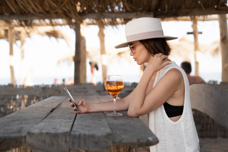 millennial woman sits in a tropical bar at a wooden table in sunglasses and a hat holds her smartphone in her hands flipping a news feed. Vacation in tropical countries.の写真素材
