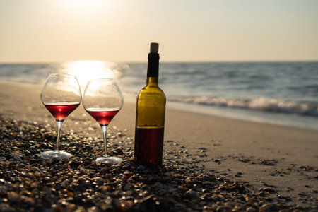 A bottle and a glass of red wine on the sand against the backdrop of the sea or ocean at sunset.