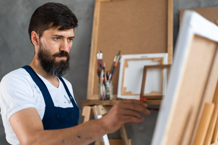 portrait of a bearded male artist in a dirty apron stands in an art studio, holding a brush in his...