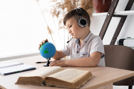 A sad schoolboy boy sitting in headphones at a desk listens to a geography lesson and looks at the...
