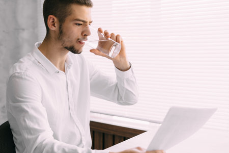 Portrait of a serious man drinking pure mineral water from a glass in the office. businessman is studying the report of the company and drinks clear water. dehydration. health concept.の写真素材