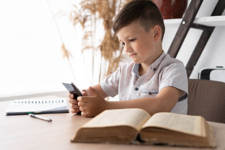 concentrated boy playing a game on the mobile phone while sitting at the table does homework. Gadget addicted student. Concept for online games or social network.の写真素材