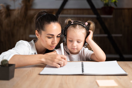 Happy young caucasian mom and handwriting small daughter in notebook prepare school home task...