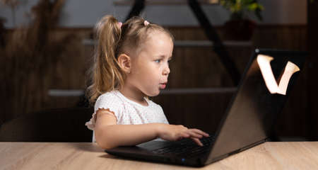A little Caucasian girl sits alone at home at a table in the evening watching a video online on a laptop. emotions of surprise on the face.の写真素材