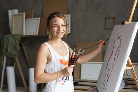 A woman artist works in an art studio stands near the canvas and paints with a brush.の写真素材