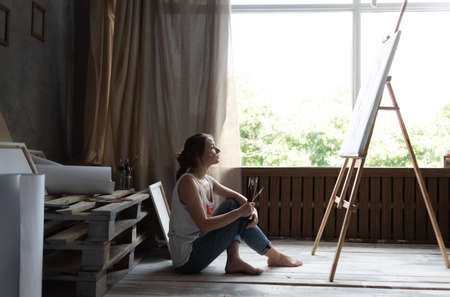in an atmospheric art studio, a beautiful Caucasian woman artist sits on the floor looking at the easel and examines the picture she has painted.の写真素材