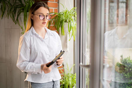 business woman standing in the office by the window with a notebook in her hands. Serious upset secretary with glasses at the panoramic window. Office worker with a paper client base. work concept.の写真素材