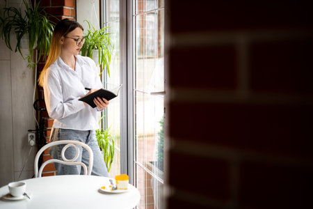 A journalist girl in glasses with a notebook in her hands standing at the window of a cafe in the...