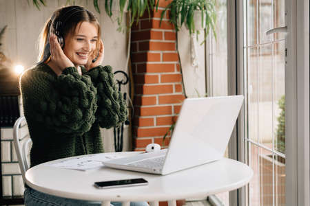 Happy girl in headphones with a laptop has a freelance job while sitting in a cafe. Cheerful woman with wireless headset listening to music or online webinar. webcam education. copy spaceの写真素材