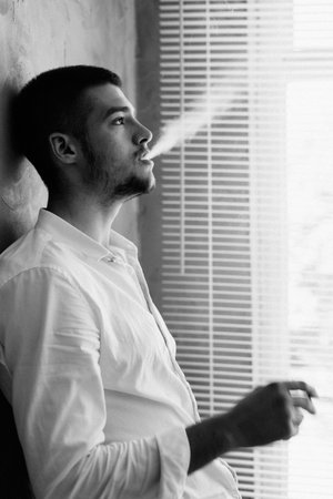 Portrait of a young man smoking a cigarette standing in the office by the window. Businessman and...