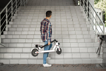 A man holds a folded electric scooter in his hand on the stairs near the building. rises up....