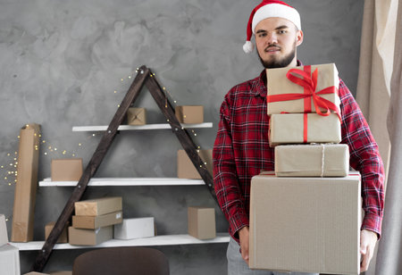 Portrait of a male small business owner in a Santa Claus hat standing in the office with cardboard boxes of gifts in his hands on Christmas Eve or New Year 2022. Delivery of ordersの写真素材