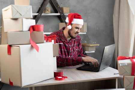 Santa Claus accepts applications on the Internet while sitting in an office with a laptop and boxes for gifts for Christmas. A man in a santa hatの写真素材