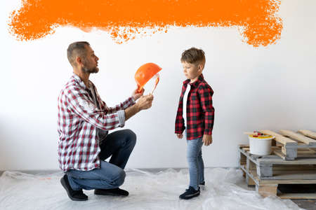 Dad and son are renovating a room in the house. A man puts on a helmet for his son against a white wall with an orange spot. Repair and painting work. Place for text.の写真素材
