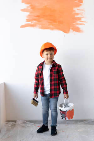 A boy wearing a shirt and jeans is standing near a white wall in an orange helmet with paint and a brush in his hands. The child painted the wall.の写真素材