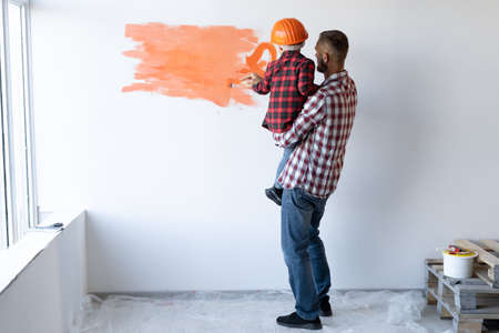 Back view of a family doing home repairs father holds his son in his arms and he paints the wall orange. A helmet on the head of a little painter.の写真素材