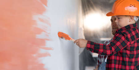 Profile view of a boy in a hard hat with dad painting the wall in the room in orange. The family is doing home repairs. Mens work and colored wall.の写真素材