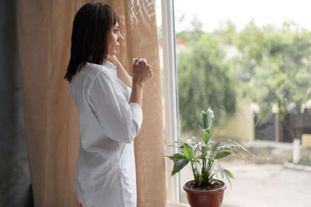 morning hot coffee in the hands of a Caucasian brunette standing in the bedroom by the window. A girl in a shirt drinks a hot drink and is resting looking outside the profile view.の写真素材