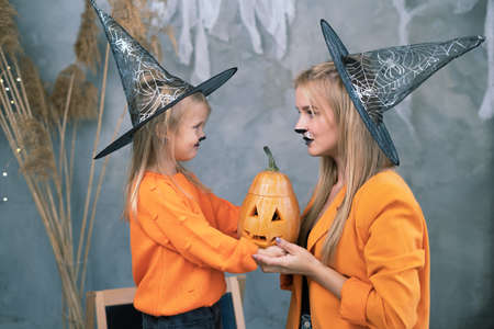 Family mom and daughter in witch hats with pumpkin jack-o-lantern celebrate Halloween at home. woman and a girl with makeup at the Halloween festivalの写真素材