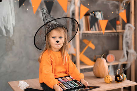 Portrait of a little girl in a Halloween costume sitting at the table with a palette of eyeshadows for makeup in a witch hat. Holiday on October 31の写真素材