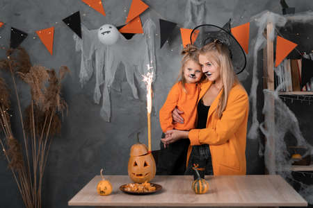 Happy mother with daughter wearing Halloween hats hold look at orange pumpkin jack-o-lantern fireworks for party in decorated room. Halloween conceptの写真素材
