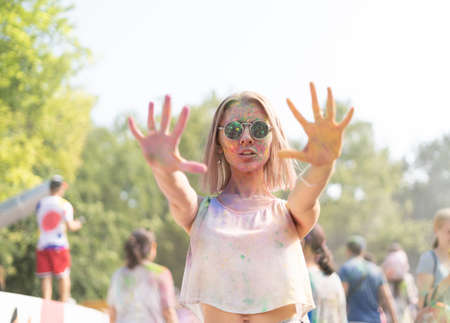 Millennial girl at the Holi Fest festival of paints in city park shows stop rocky. Traditional festival of dry urban paints. woman in glasses with face stained with paintsの写真素材