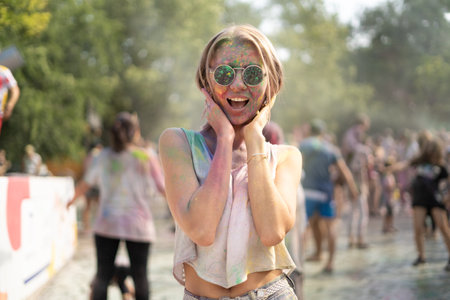 Blonde girl at the Color Fest festival of paints in the city park. Traditional festival of dry paints in the city. Surprised woman in glasses with face paintedの写真素材