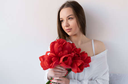 Portrait of an attractive girl with a bouquet of red tulips in the morning in the bedroom for Happy Birthday. woman with flowers on the bed close-up.の写真素材