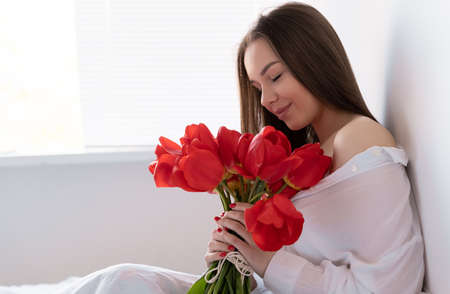Portrait of a sexy girl with a bouquet of red tulips in the morning in the bedroom for Valentines Day. Woman with flowers on the bed Womens dayの写真素材