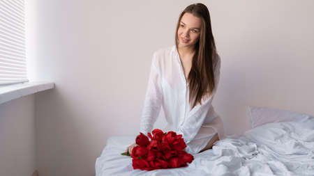 Sexy girl in the bedroom on the bed with a bouquet of flowers of red tulips sits happy on her birthday or Valentines Day. Flowers in the morningの写真素材