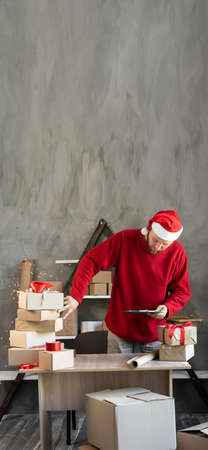 A man in a Santa hat works in a warehouse counting boxes with gifts and goods from an online store. Small business home office and Christmas saleの写真素材