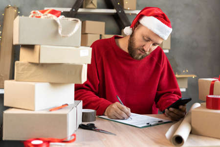 male entrepreneur business owner working at home on christmas eve wearing a red santa claus hat and sweater received a phone and writes delivery.の写真素材