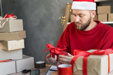 Portrait of a businessman in a Santa hat working in a home office or warehouse with boxes of gifts for Christmas. A man puts box in a shopping basketの写真素材