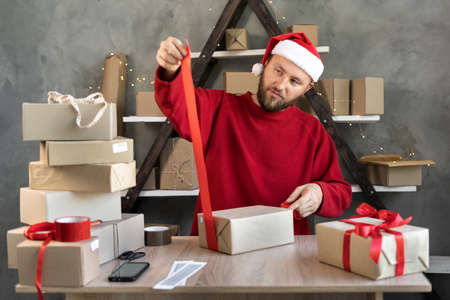 a man in a Santa hat prepares Christmas gifts in a warehouse for delivery and ties a red ribbon. Cardboard boxes on the table. New Year saleの写真素材